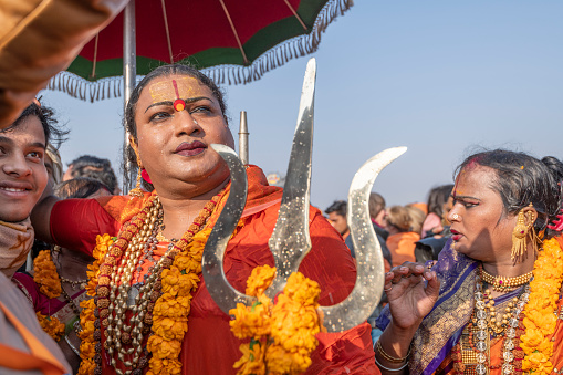 Overweight, middle-aged hijra, rearing a dark orange shirt, necklaces and yellow garlands, holds a brass trishula (trident), while coming from the holy dip, among a crowd of other hijras and pilgrims. Photo taken during Kumbh Mela 2019 in Prayagraj (Allahabad), India. Hijra are eunuchs, intersex and transgender people. They were gathered in an organization (Kinnar Akhada) and for the first time, they were allowed to set their own camp in the tent city, and had a major role on the processions, hence their joy and expressions of triumph.\nKumbh Mela or Kumbha Mela is a major pilgrimage and festival in Hinduism, and probably the greatest religious festival in the World. It is celebrated in a cycle of approximately 12 years at four river-bank pilgrimage sites: the Allahabad (Ganges-Yamuna Sarasvati rivers confluence), Haridwar (Ganges), Nashik (Godavari), and Ujjain (Shipra). The festival is marked by a ritual dip in the waters, but it is also a celebration of community commerce with numerous fairs, education, religious discourses by saints, mass feedings of monks or the poor, and entertainment spectacles. Pilgrims believe that bathing in these rivers is a means to cleanse them of their sins and favour a better next incarnation.
