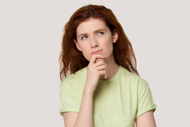 istock Doubtful unsure young red-haired woman touching chin, looking aside up. 1254296739