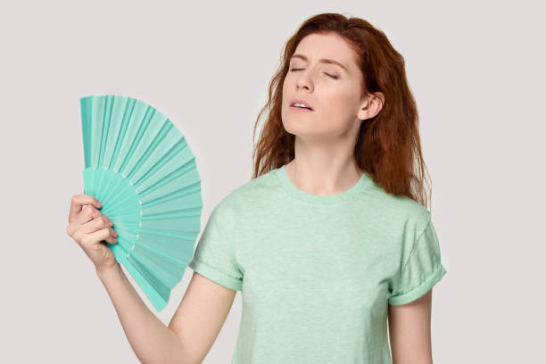 istock Overheated young red-haired woman using fan cooling herself. 1254296754