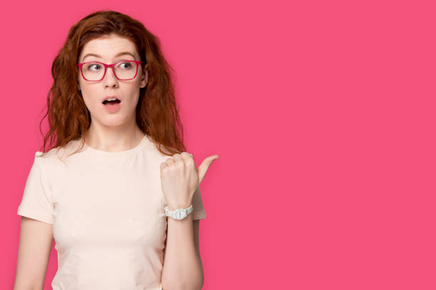 istock Surprised young red-haired woman in eyeglasses pointing finger at copyspace. 1254296762