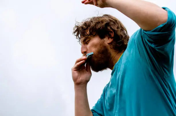 Handsome Man dancing and playing mouth organ outdoors against cloudy sky on Mountain peak. Young Man celebrating successful climbing. Copy space