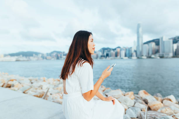 istock Confidence young Asian woman using a smartphone and sitting on a boardwalk beside the seaside with the iconic Hong Kong skyline in the background 1254320437