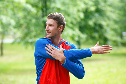 Front view of a sportsman stretching hands while standing outdoor in a park and looking by side. The concept of an active and healthy lifestyle
