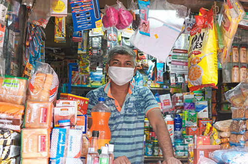 Kolkata, India, 07/05/2020: A middle aged shop owner, wearing facemask inside his stationery shop. Stacks of packaged food and other fmcg products are seen at store front. During Unlock 3.0, due to easing lockdown restrictions, more number of shops are gradually opening.