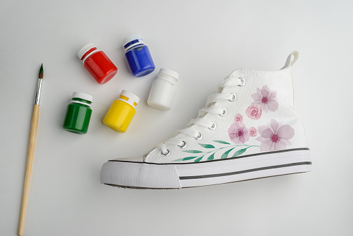 Canvas sneakers with handpainted flowers. Vials of fabric paint and brush on white background. Cute refashion project concept.