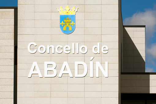Abadín, Lugo, Spain-September 30, 2019: Detail of Town hall in Abadín, Lugo province, Galicia, Spain. Text and coat of arms on the facade of a modern stone Local Government  building and clear blue sky background.