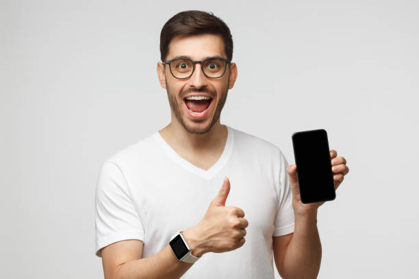 opgewekte knappe mens die leeg scherm van telefoon houdt en duim omhoog toont als als gebaar aan nieuwe app, die op grijze achtergrond wordt geïsoleerd - phone hand thumb stockfoto's en -beelden