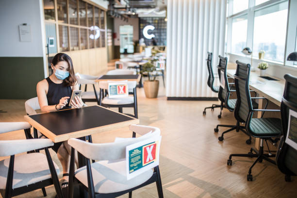 an asian chinese business woma using mobile phone wearing protective masks sitting on office chair while maintaining social distance to protect from covid-19 - ding imagens e fotografias de stock