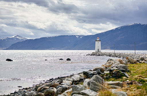 A small white lighthouse on the coastline of Norway. A path of stones leads you to it. The sky is filled with clouds and in the background are mountains.