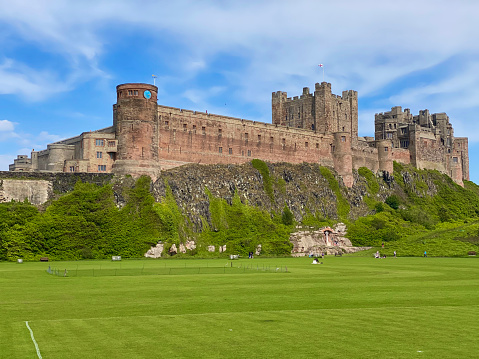 Bamburgh, England, United Kingdom – June 20, 2020: Bamburgh Castle is a medieval castle located on the coast of the North Sea along Bamburgh Beach in the village of Bamburgh. Today the castle is privately owned by the Armstrong family but is open to the public for sightseeing and events. Here, we can see people sunbathing and picnicking in the open sports field by the castle.