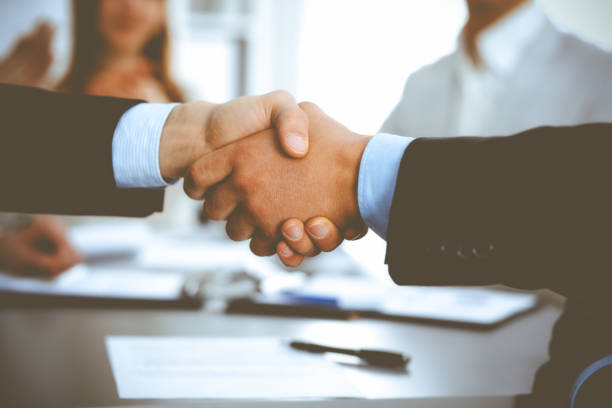 istock Business people shaking hands at meeting while theirs colleagues clapping and applauding. Group of unknown businessmen and women in modern white office. Success teamwork, partnership and handshake concept 1254407165