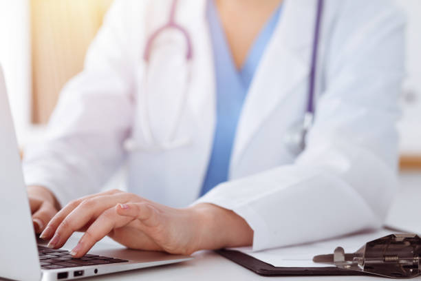 istock Unknown woman-doctor typing on laptop computer while sitting at the table in sunny clinic 1254409351
