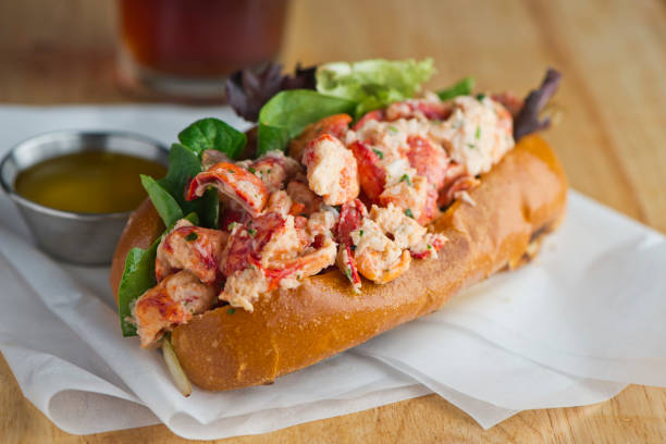Lobster roll. Maine lobster mixed with mayo, celery, onions, garlic, scallions, chives, lemon juice. Lobster roll on toasted hotdog bun w/ lettuce, tomato, garlic mayo seasoned salt & pepper. Classic American restaurant or diner lunch sandwich favorite. rollo de langosta. langosta de maine mezclada con mayonesa, apio, cebolla, ajo, cebolletas, cebollino, jugo de limón. rollo de langosta en bollo de perrito caliente tostado con lechuga, tomate, mayonesa de ajo sal sazonada y pimienta. clásico restaurante