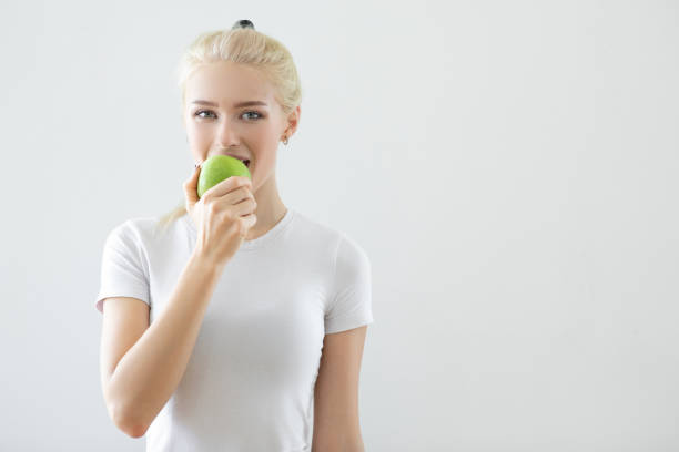 Stylish young woman in white and plaid. Squatting to smell a green apple. Exuding desire and beauty with red heels. Embodying a healthy lifestyle. Captivating studio shot of modern. Trendy elegance
