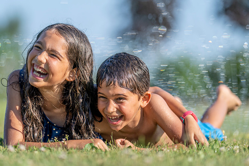 Hispanic brother and sister playing in a water sprinkler during the summertime