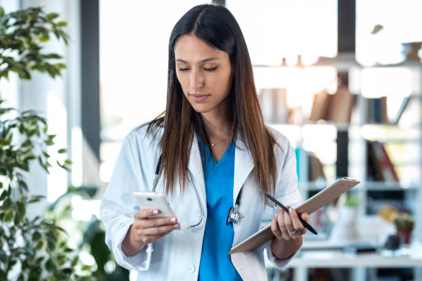 mujer doctora segura usando su teléfono móvil mientras sostiene un portapapeles y está de pie en la consulta. - medical texts fotografías e imágenes de stock