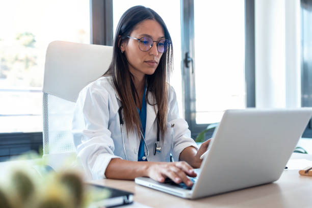 Confident young female doctor using her mobile phone while working on laptop in the consultation. Shot of confident young female doctor using her mobile phone while working on laptop in the consultation. physician computer stock pictures, royalty-free photos & images