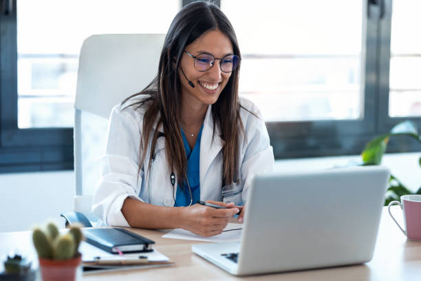 Female doctor talking with colleagues through a video call with a laptop in the consultation. Shot of female doctor talking with colleagues through a video call with a laptop in the consultation. remote control stock pictures, royalty-free photos & images