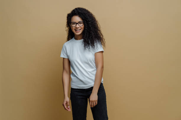 istock Smiling dark skinned young woman with glad face expression, wears optical glasses, white t shirt and jeans, poses carefree against beige background. People, happy mood and lifestyle concept. 1254447936