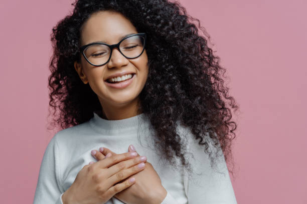 primo scatto di felice donna afro-americana sincera tiene le mani premute sul petto, esprime sentimenti sinceri, fa gesto di gratitudine, si sente grata di aiuto, sorride in generale, indossa occhiali - fashion male beauty confidence elegance foto e immagini stock