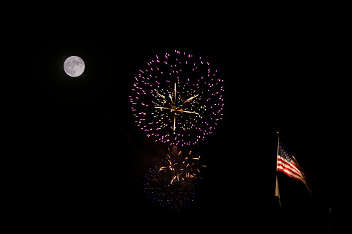 This photo was taken during a full moon and shows a firework display along with an American flag flying on a pole.  The shot was taken from a distance so that the moon, fireworks, and flag are all in focus and in frame.  Illumination levels of the moon and fireworks were seperately adjusted so that details of both are clear.