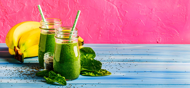 Green smoothies in glass bottles on cool pink blue background with yellow bananas. Panoramic banner with copy space