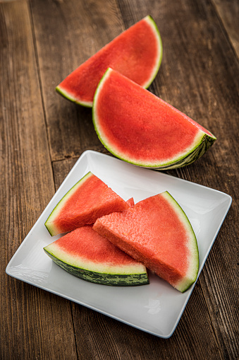 This is an outdoor photograph of sliced red see this watermelon on a white square modern plate sitting on a wooden picnic bench outdoors for a simple and concept of healthy eating and snacks during the summertime fun.