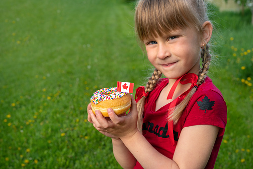 Happy Canada Day Celebration Concept. Young Cute Caucasian Girl smiling looking to the camera and holding sprinkled donut with Canadian flag toothpick. Copy Space on right. National Day. 1st of July.