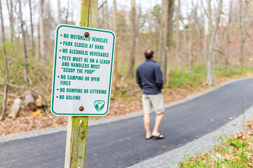 Herndon, USA - March 14, 2020: Rules information sign during spring in Fairfax County Northern Virginia on Sugarland Run Stream Valley Trail with paved road path in forest and man