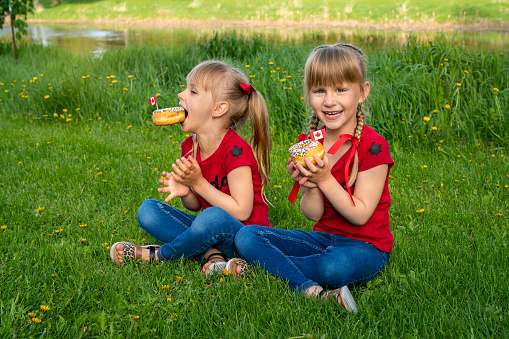 Happy Canada Day Celebration Concept. Young Cute Caucasian Girls Sisters smiling and eating donuts decorated with Canadian flag toothpick sitting on grass in park outside. Canada Day Anniversary.