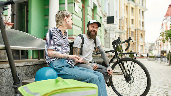 Two cheerful couriers, young man and woman sitting on the bench and talking outdoors while delivering food and products, using scooter and bike. Courier, delivery service concept. Focus on people