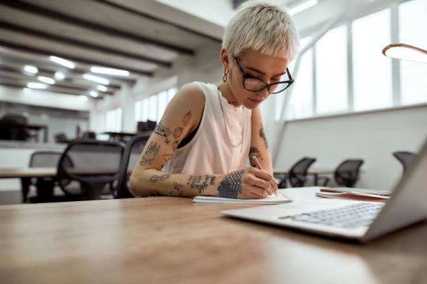 istock Planning the day. Young, beautiful tattooed business lady in eyewear making some notes while sitting at her desk in the modern office 1254502120