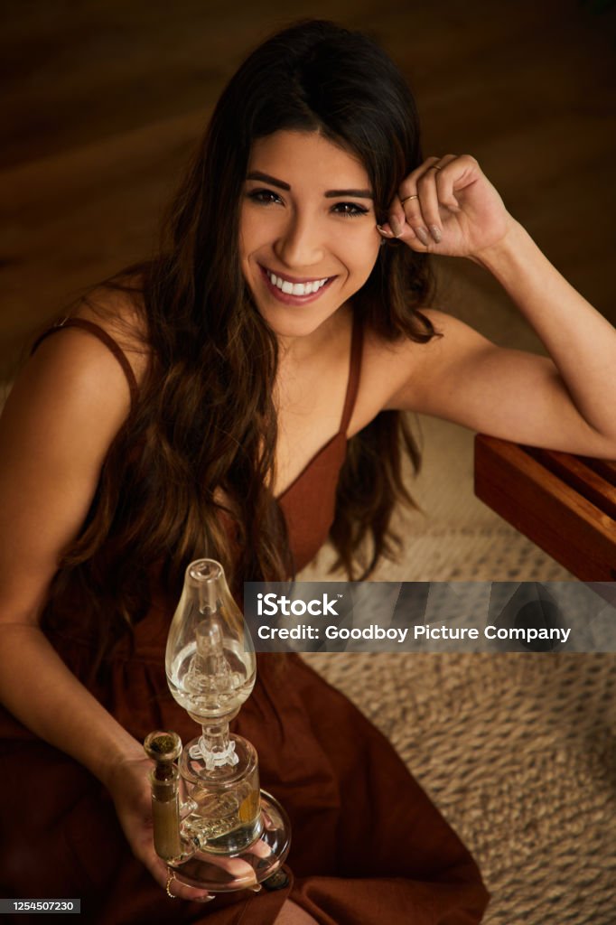 Smiling young woman sitting at home holding a glass bong Portrait of a smiling young woman holding a glass bong while sitting on her living room floor at home Bong Stock Photo