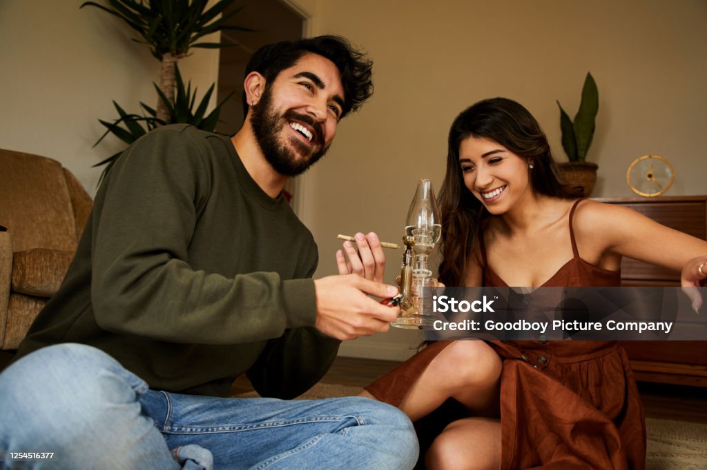 Young couple sitting at home smoking a bong and laughing Young couple laughing while sitting together on their living room floor at home and smoking weed in a bong Bong Stock Photo