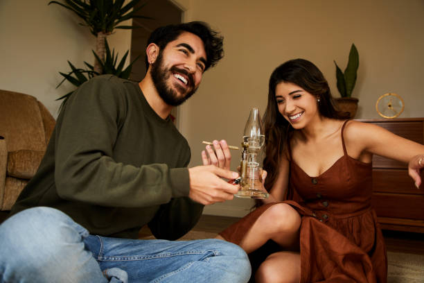 Young couple sitting at home smoking a bong and laughing Young couple laughing while sitting together on their living room floor at home and smoking weed in a bong bong stock pictures, royalty-free photos & images