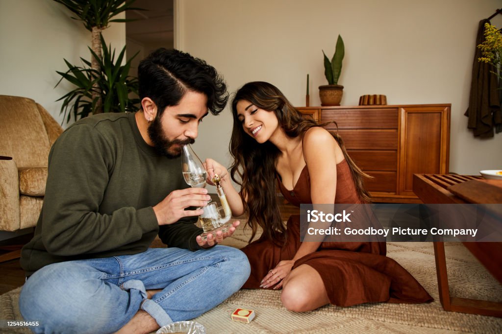 Smiling young couple smoking a bong Smiling young couple sitting on their living room floor at home and smoking weed together in a bong Bong Stock Photo