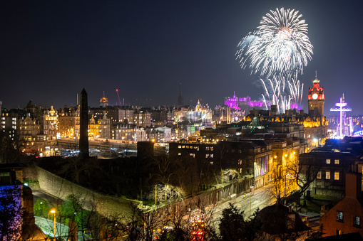 View from the top of Calton Hill in Edinburgh, Scotland. Looking across at bright fireworks exploding over the city on New Year's Eve. The colourful display is set off to welcome in the new year. Crowds of people gather in the streets below and on surrounding hills to watch the display. Edinburgh castle can be seen in the background lit up with purple lighting. The tall fun fair structures of the Christmas Market can be seen in Princes Street below.