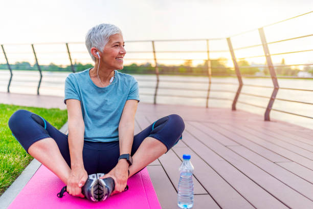 Woman on a yoga mat to relax outdoor. Senior woman exercising in park while listening to music. Senior woman doing her stretches outdoor. Athletic mature woman stretching after a good workout session. exercising stock pictures, royalty-free photos & images