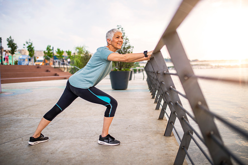 Smiling retired woman stretching legs outdoors. Senior woman enjoying daily routine warming up before running at morning. Sporty lady doing leg stretches before workout