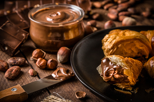 High angle view of some croissants smeared with chocolate and hazelnut spread surrounded by a bread knife, a crystal jar full of chocolate and hazelnut spread and some whole and peeled hazelnuts on a rustic wooden table. Main focus is on the croissants and at the defocused background are some chocolate bars and cocoa beans. Predominant color is brown. Low key DSLR photo taken with Canon EOS 6D Mark II and Canon EF 24-105 mm f/4L