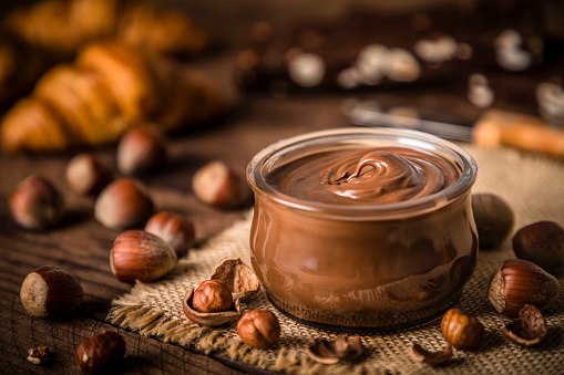Front view of a crystal jar full of chocolate and hazelnut spread surrounded by some hazelnuts on a rustic wooden table. Selective focus is on the jar and on the defocused background are some croissants and chocolate bars. Predominant color is brown. Low key DSLR photo taken with Canon EOS 6D Mark II and Canon EF 24-105 mm f/4L