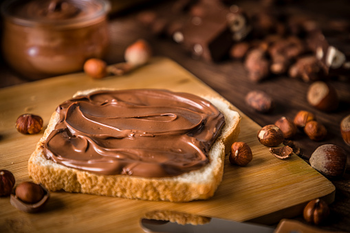 High angle view of a wooden cutting board with a slice of bread full of chocolate and hazelnut spread on top surrounded by some whole and peeled hazelnuts, some cocoa beans and a crystal jar full of chocolate and hazelnut spread. Predominant color is brown. Low key DSLR photo taken with Canon EOS 6D Mark II and Canon EF 24-105 mm f/4L