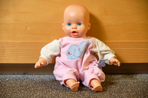 Portrait of an old used toy doll representing a caucasian baby sitting in front of a bright wall of a cabinet on the carpeted floor