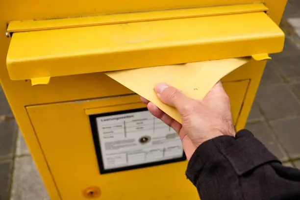 Man's hand putting an envelope into the mail slot of a yellow letterbox in Germany in December.