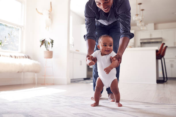 padre animando a la hija sonriente a dar los primeros pasos y caminar en casa - bebé fotografías e imágenes de stock