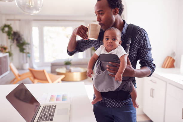 Father With Baby Daughter In Sling Multi-tasking Working From Home On Laptop Father With Baby Daughter In Sling Multi-tasking Working From Home On Laptop baby carrier stock pictures, royalty-free photos & images