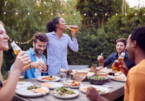 amici multiculturali a casa intorno al tavolo che bevono birra mentre si godono il cibo alla festa in giardino estivo - festa in giardino foto e immagini stock