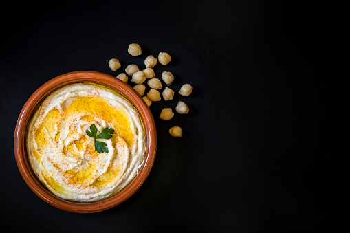 Top view of a homemade hummus with chick peas seasoned with paprika and olive oil. The plate with hummus is at the lower left corner leaving a useful copy space at the right side on a black background. Low key DSLR photo taken with Canon EOS 6D Mark II and Canon EF 24-105 mm f/4L