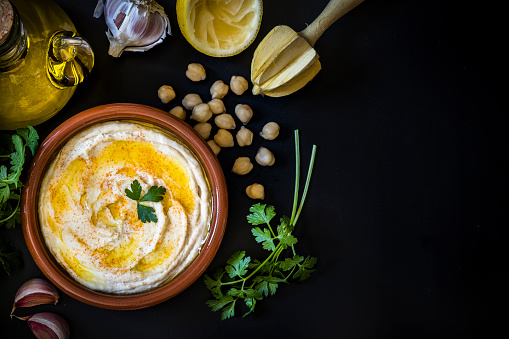 Top view of a homemade hummus with chick peas seasoned with paprika and olive oil. The plate with hummus is surrounded by an olive oil bottle, garlic cloves, some coriander leaves and some chick peas and are at the left side of the image leaving a useful copy space at the right side on a black background. Low key DSLR photo taken with Canon EOS 6D Mark II and Canon EF 24-105 mm f/4L