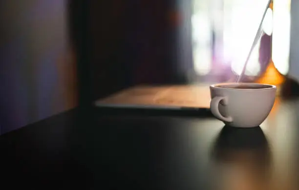 Close-up white coffee cup on desk and blurred laptop on background, Dark tone and color lighting